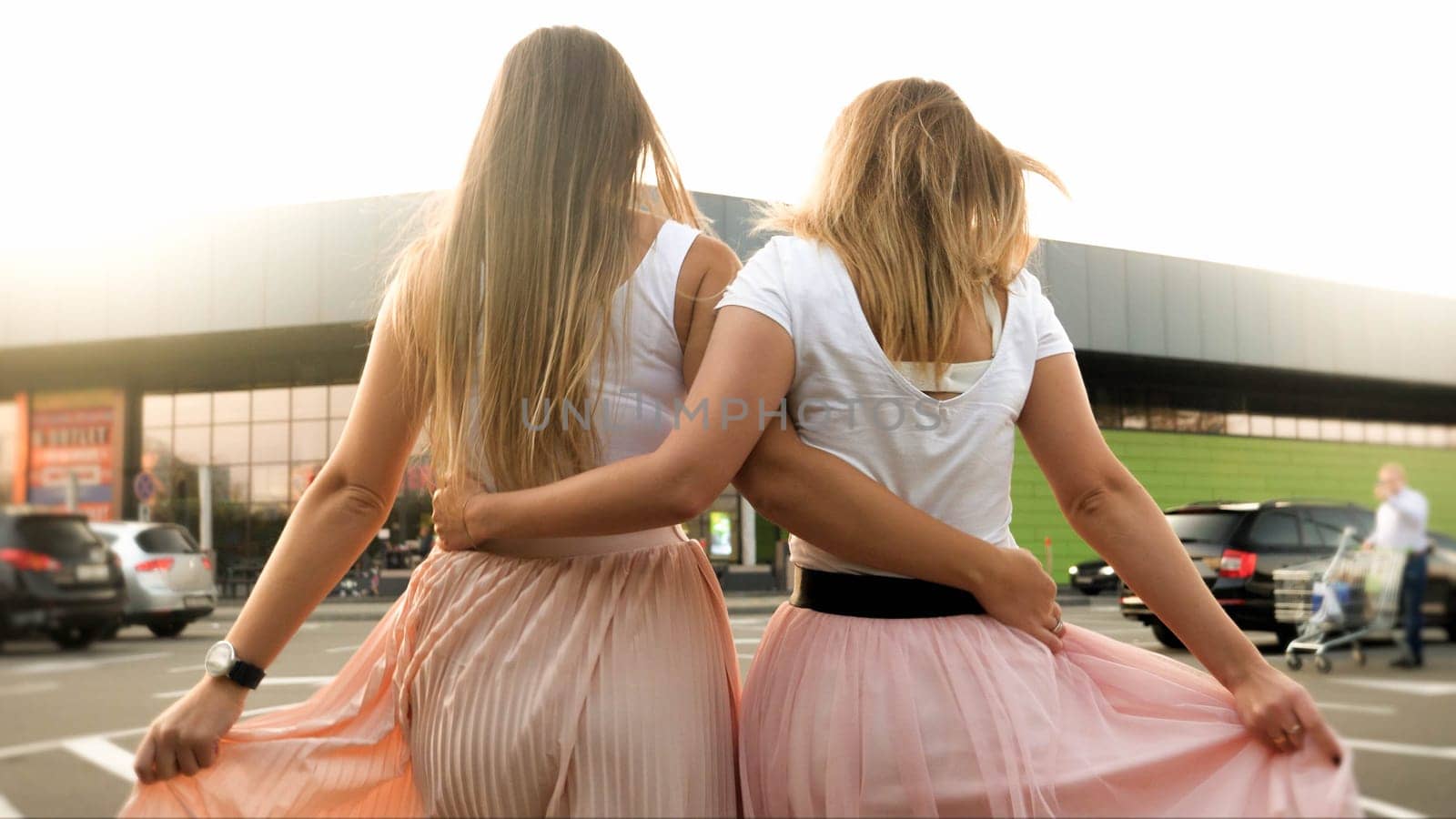 Rear view image of two beautiful girls in pink skirt embrace and holding each other while walking on car parking at shopping mall.