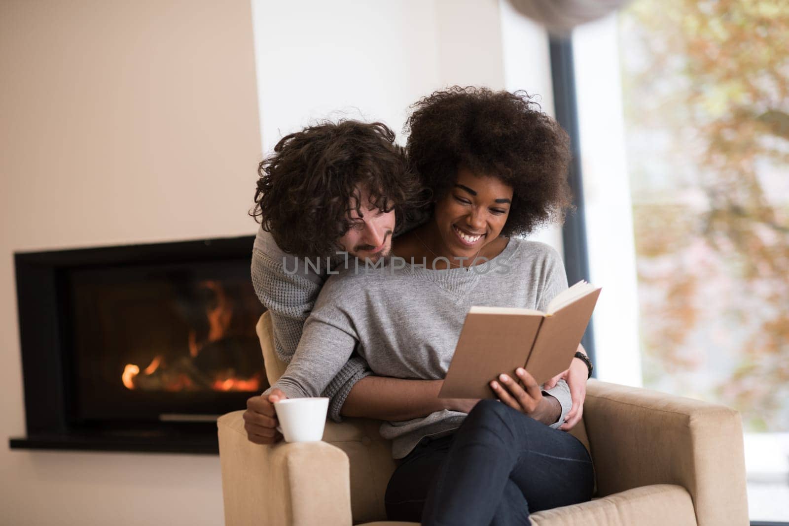 Young beautiful multiethnic couple hugging in front of fireplace at home when reading a book at autumn day