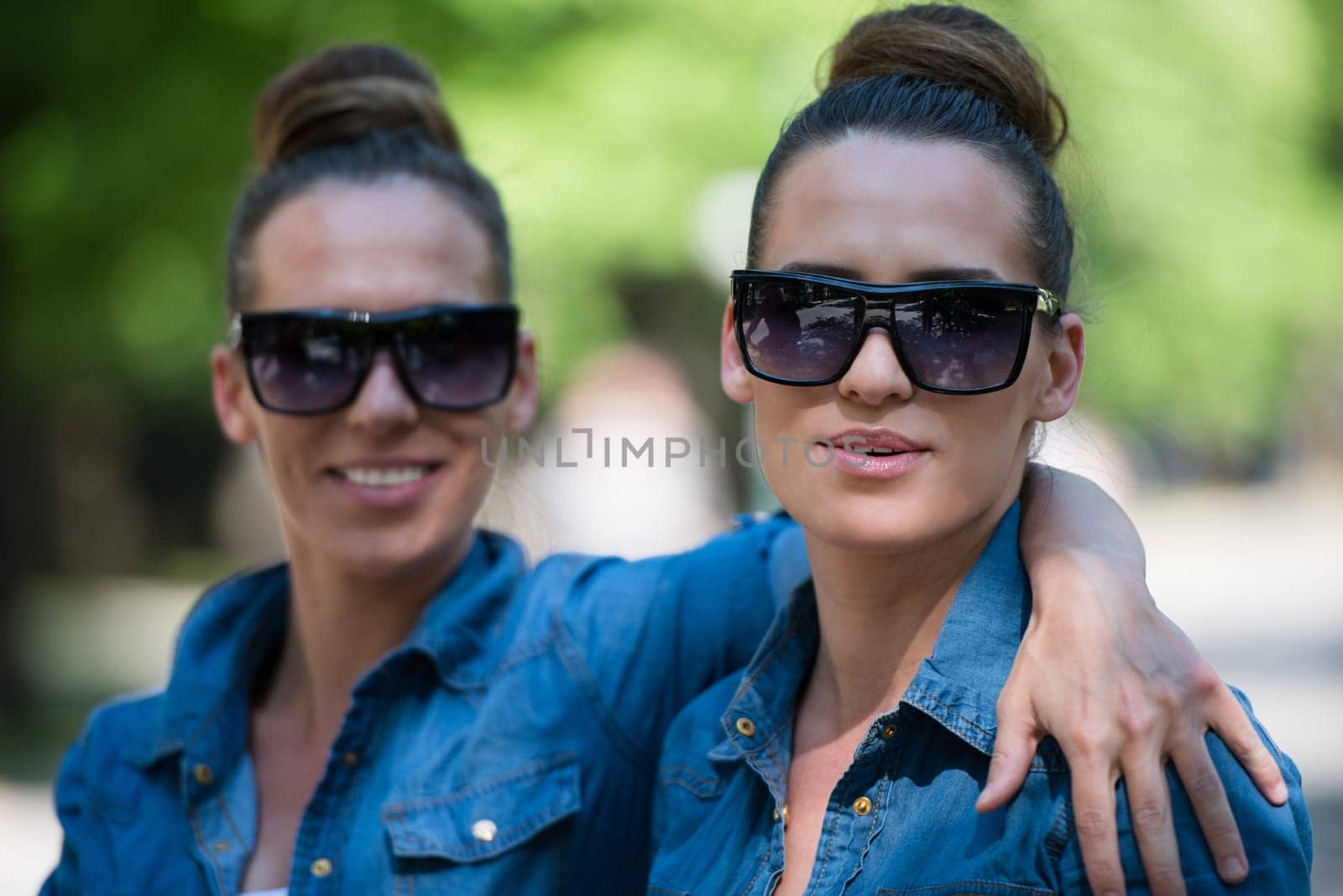 Young beautiful twin sister with sunglasses in identical wardrobe posing in a park on a sunny summer day
