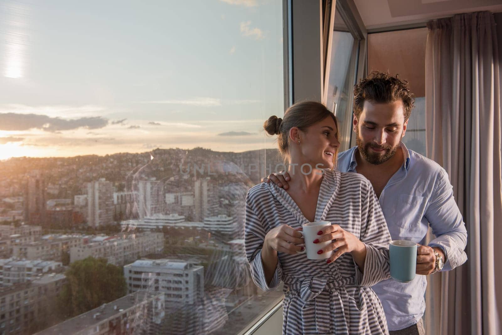 romantic happy young couple enjoying evening coffee and beautiful sunset landscape of the city while standing by the window