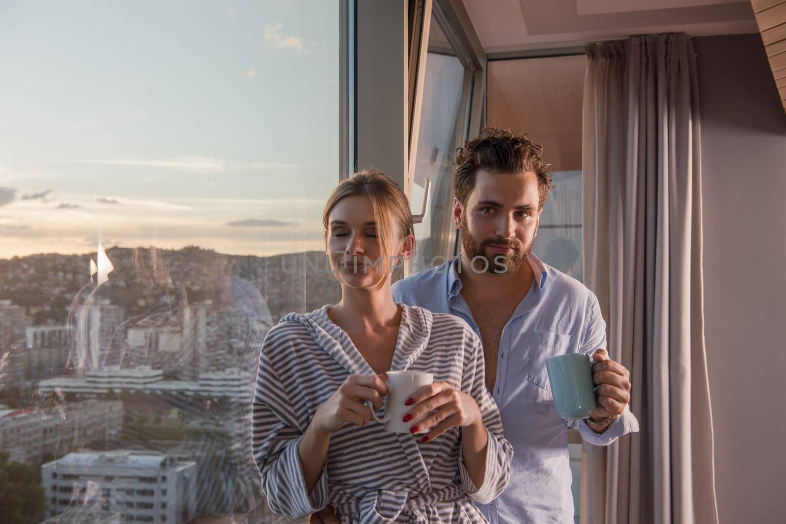 romantic happy young couple enjoying evening coffee and beautiful sunset landscape of the city while standing by the window