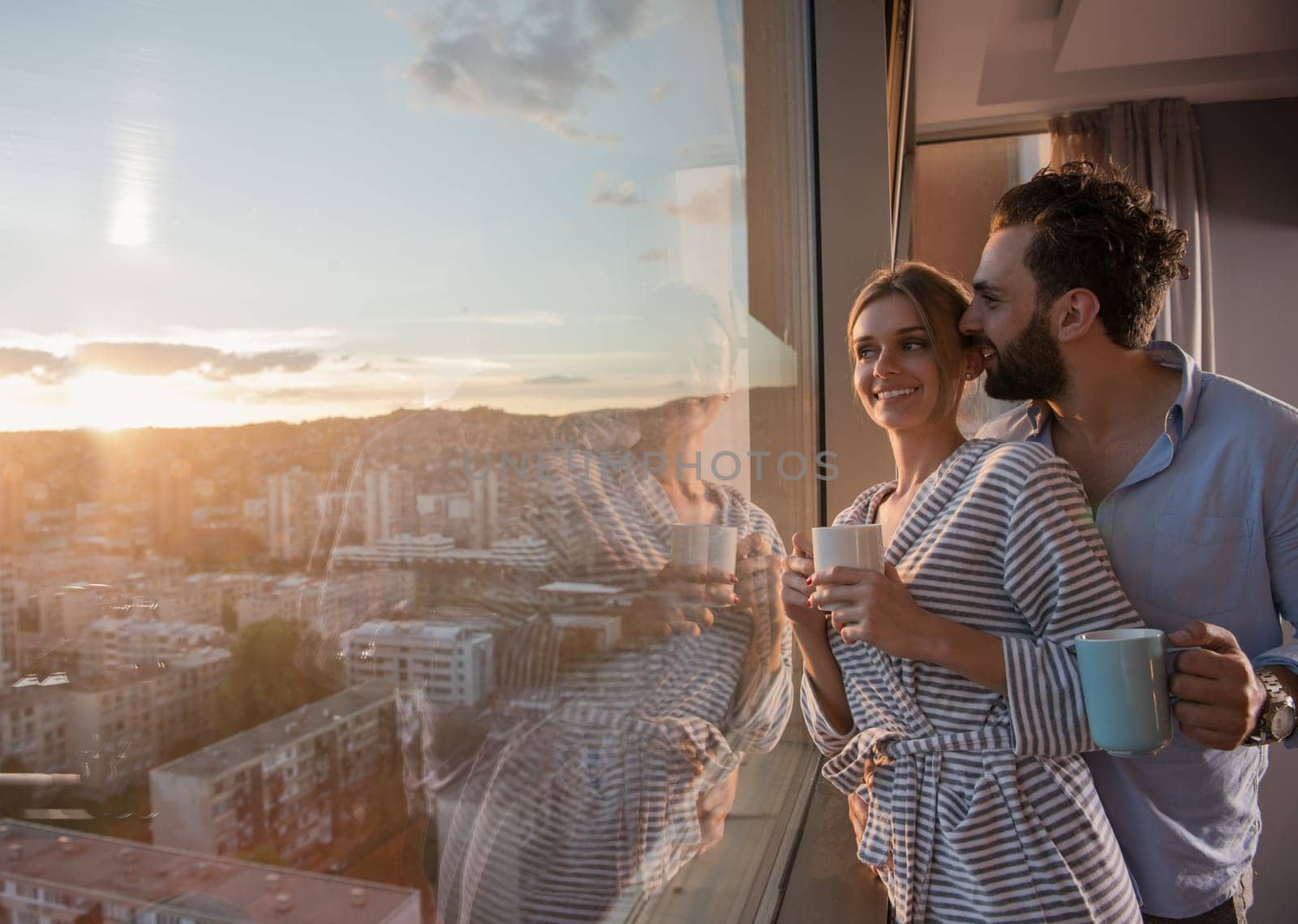romantic happy young couple enjoying evening coffee and beautiful sunset landscape of the city while standing by the window