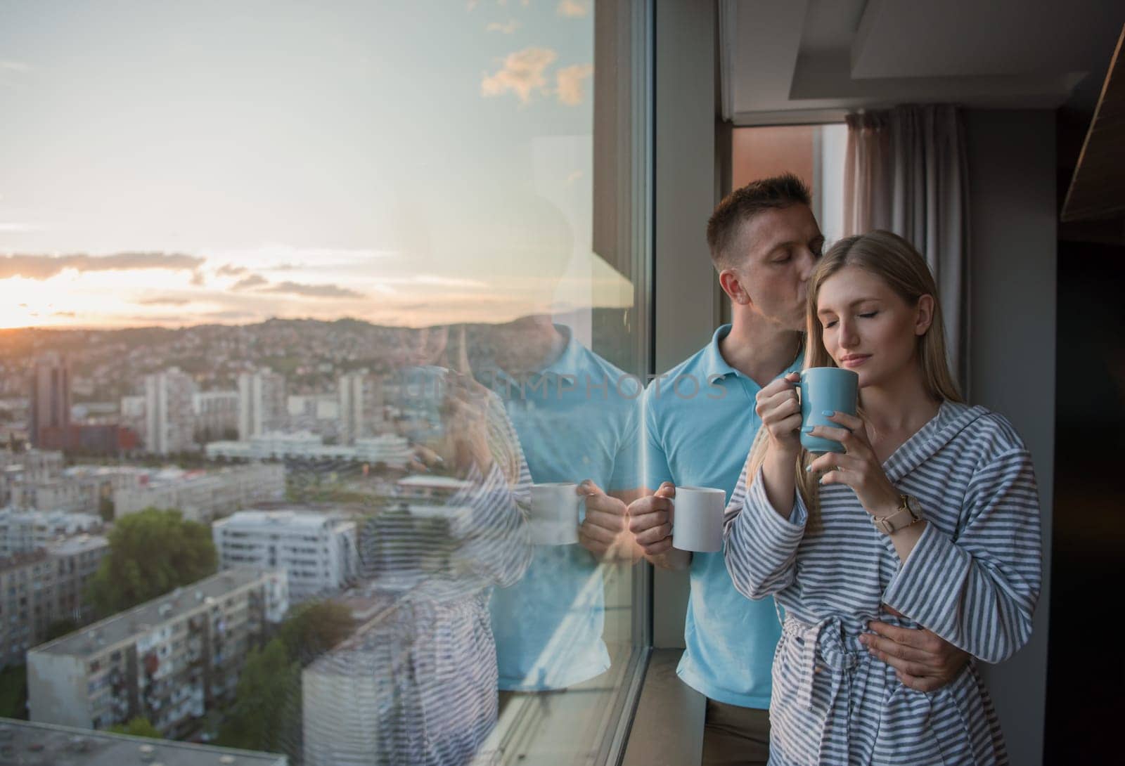 romantic happy young couple enjoying evening coffee and beautiful sunset landscape of the city while standing by the window