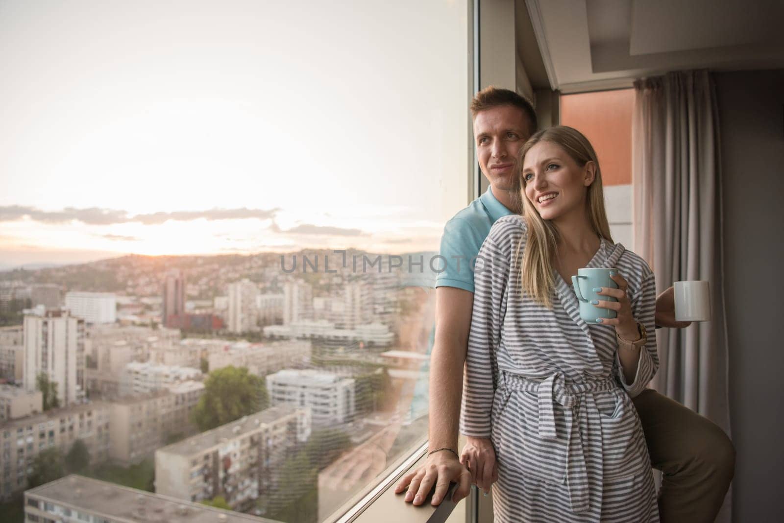 romantic happy young couple enjoying evening coffee and beautiful sunset landscape of the city while standing by the window