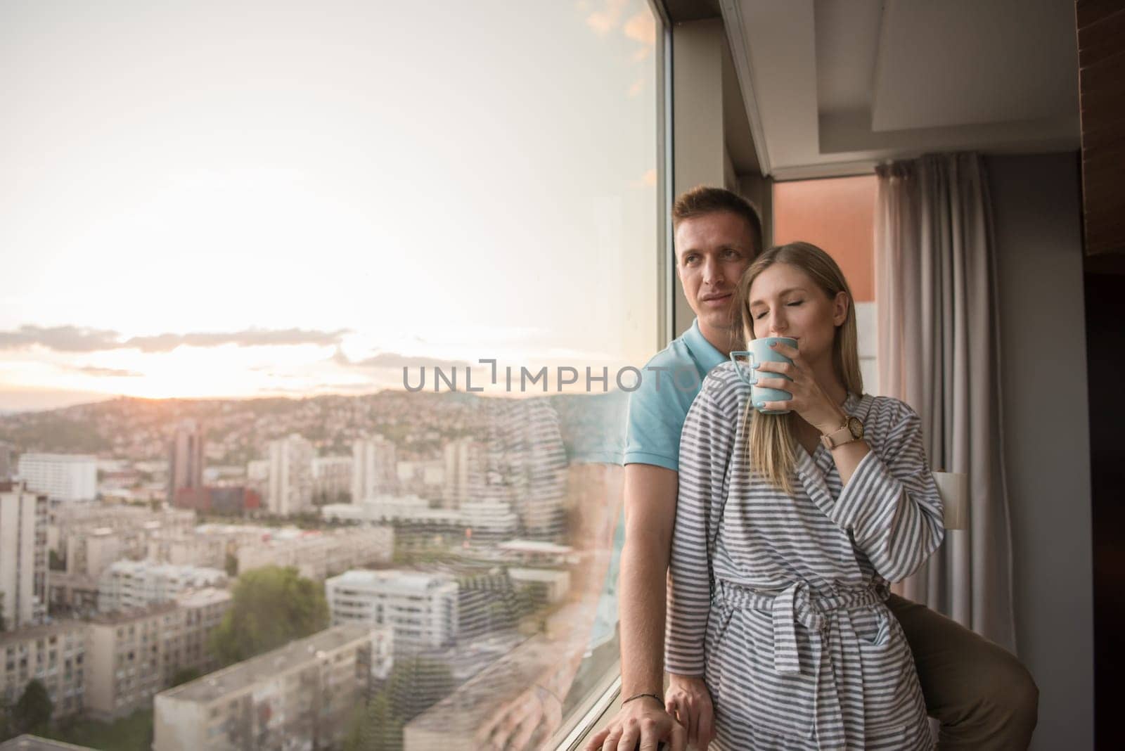 romantic happy young couple enjoying evening coffee and beautiful sunset landscape of the city while standing by the window