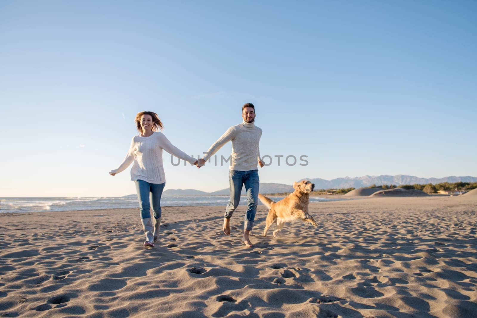 Couple Running On The Beach Holding Their Hands with dog On autmun day