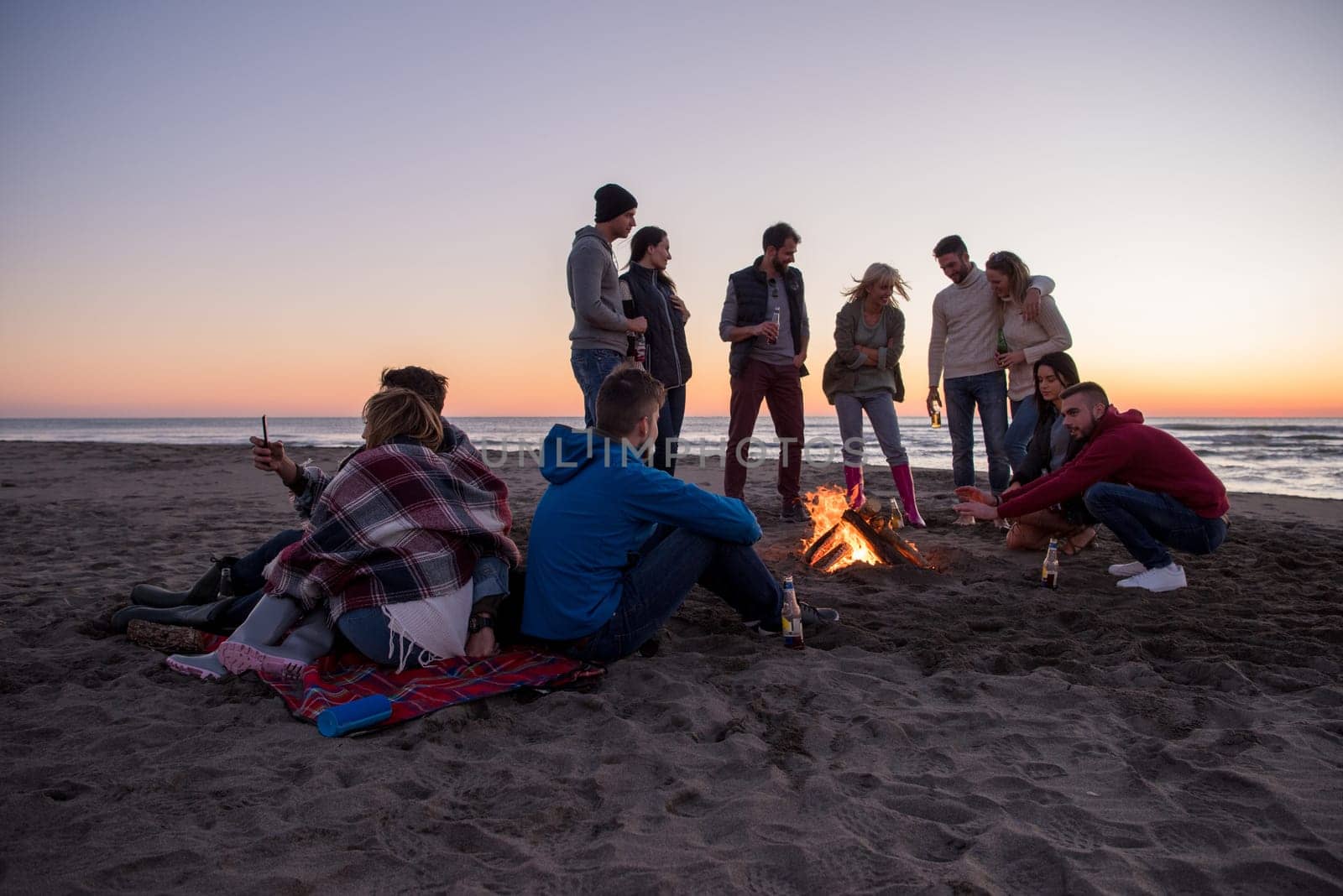 Happy Carefree Young Friends Having Fun And Drinking Beer By Bonefire On The Beach As The Sun Begins To Set