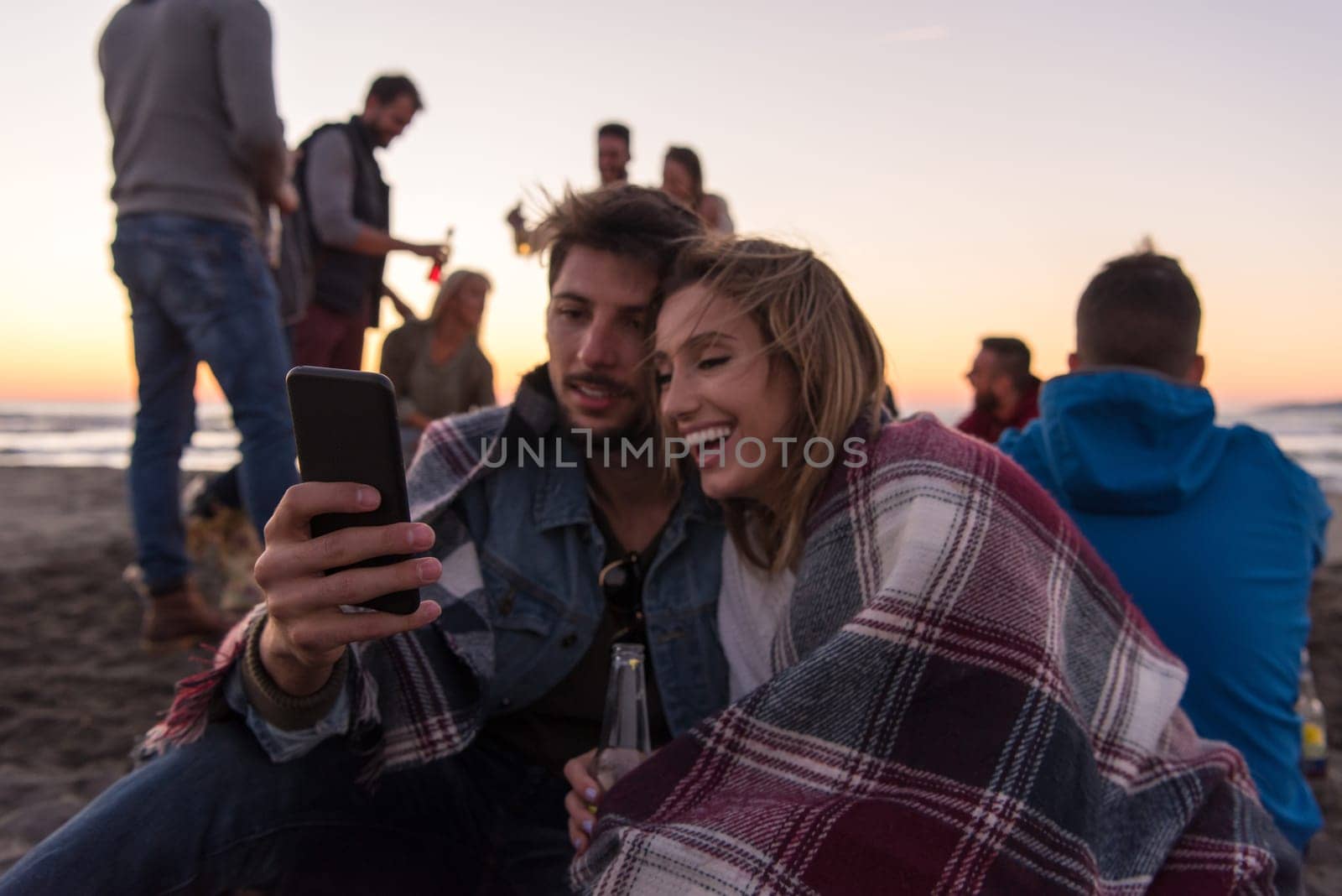 Couple using cell phone during autumn beach party with friends drinking beer and having fun