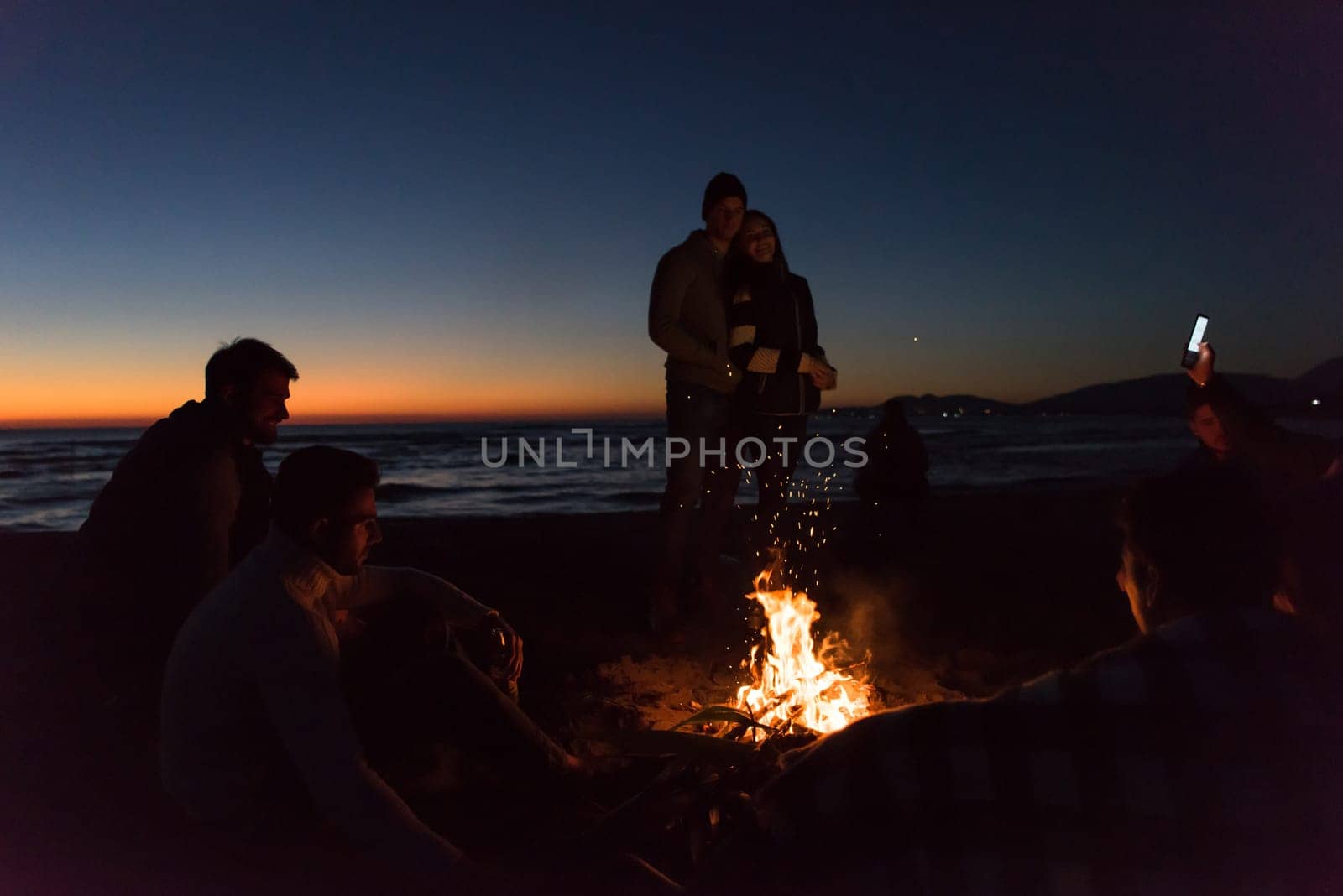 Happy Carefree Young Friends Having Fun And Drinking Beer By Bonefire On The Beach As The Sun Begins To Set