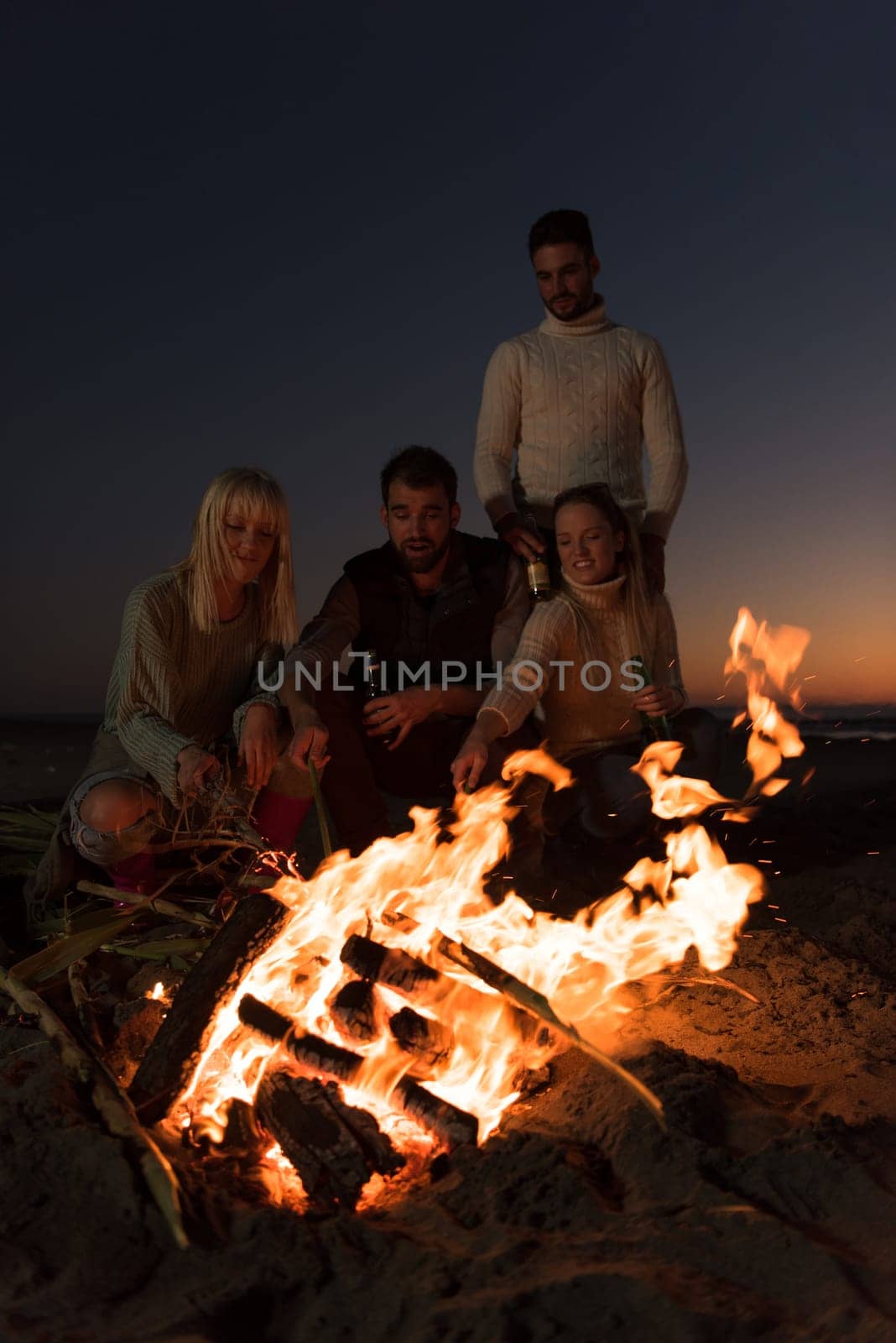 Happy Carefree Young Friends Having Fun And Drinking Beer By Bonefire On The Beach As The Sun Begins To Set