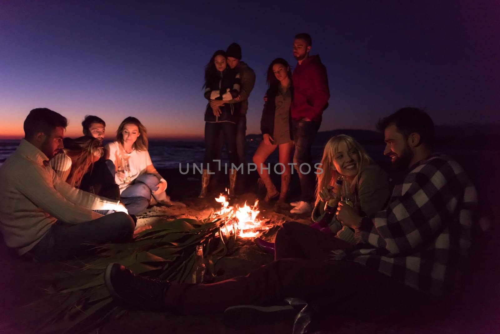 Young Couple enjoying with friends Around Campfire on The Beach At sunset drinking beer