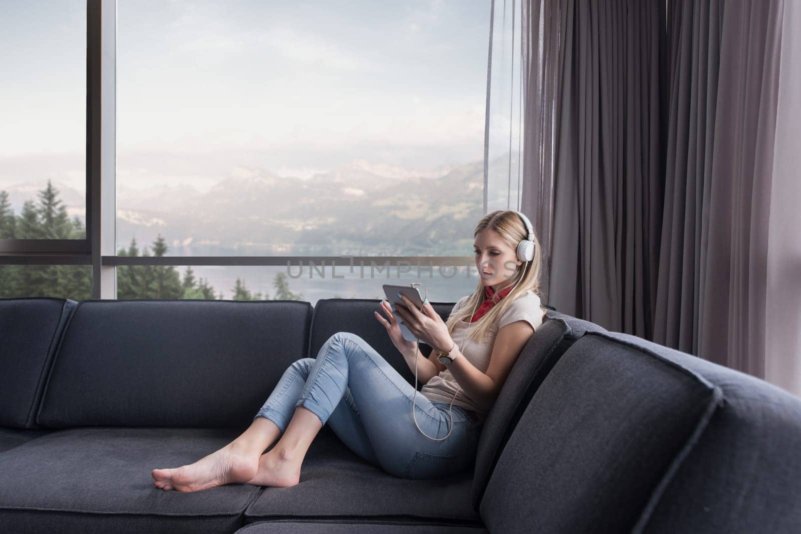 Young beautiful girl enjoying music through headphones, laying on sofa near the window at home