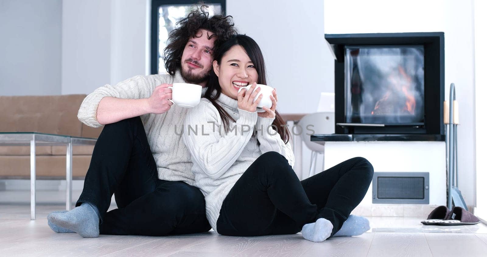 Young romantic multiethnic couple sitting on the floor in front of fireplace at home, looking at each other, talking and drinking coffee at autumn day