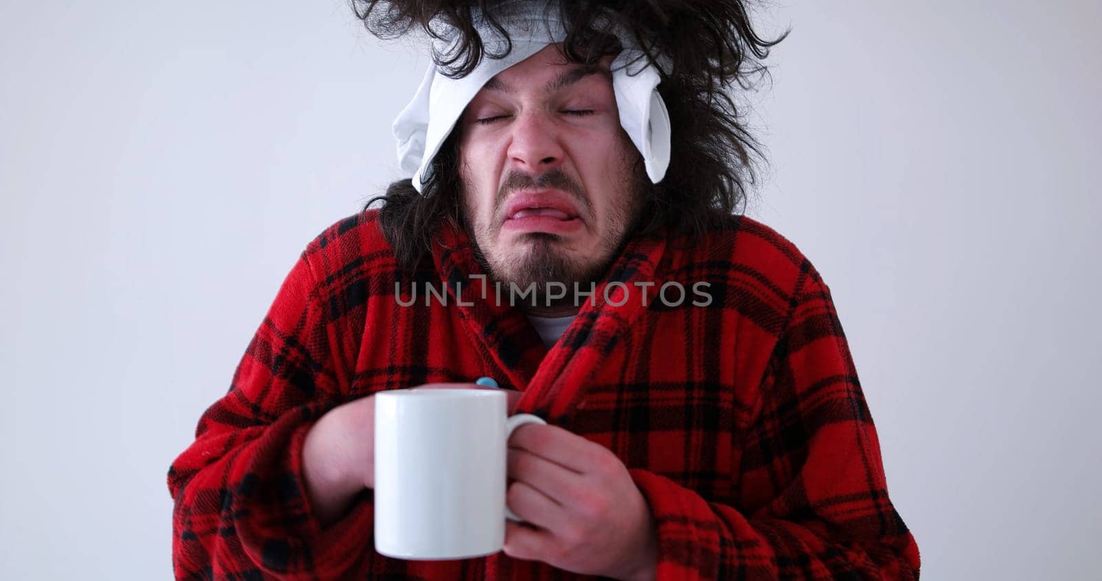 young Man with flu and fever wrapped holding cup of healing tea isolated over white