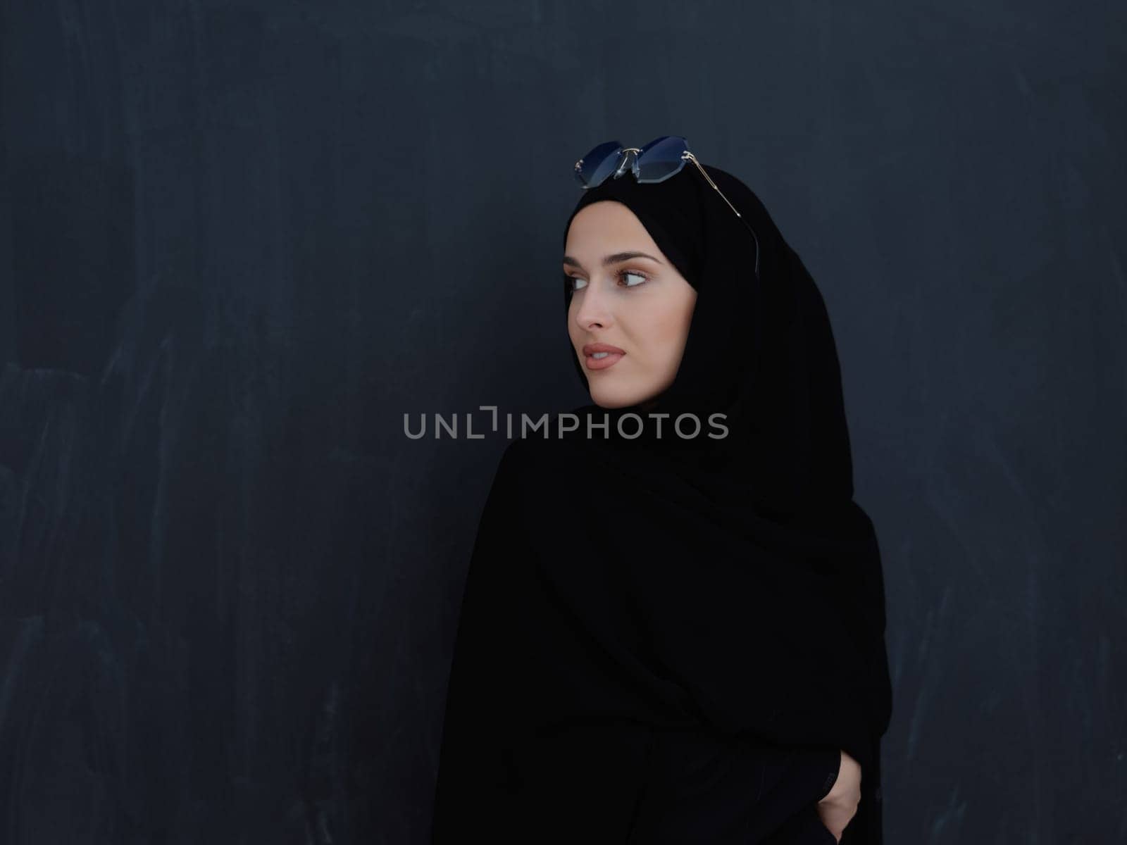 Young muslim in traditional clothes or abaya and sunglasses posing in front of black chalkboard. Arab woman representing modern arabic lifestyle, islamic fashion and Ramadan kareem concept