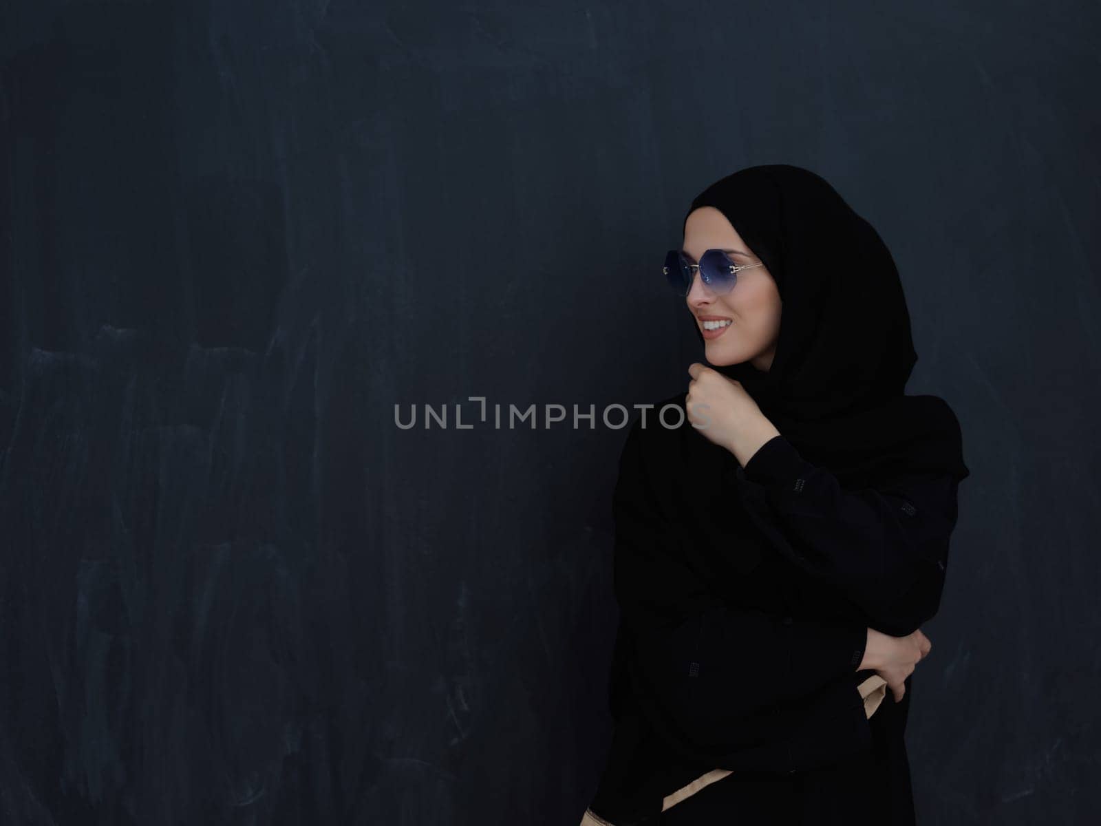 Young muslim in traditional clothes or abaya and sunglasses posing in front of black chalkboard. Arab woman representing modern arabic lifestyle, islamic fashion and Ramadan kareem concept