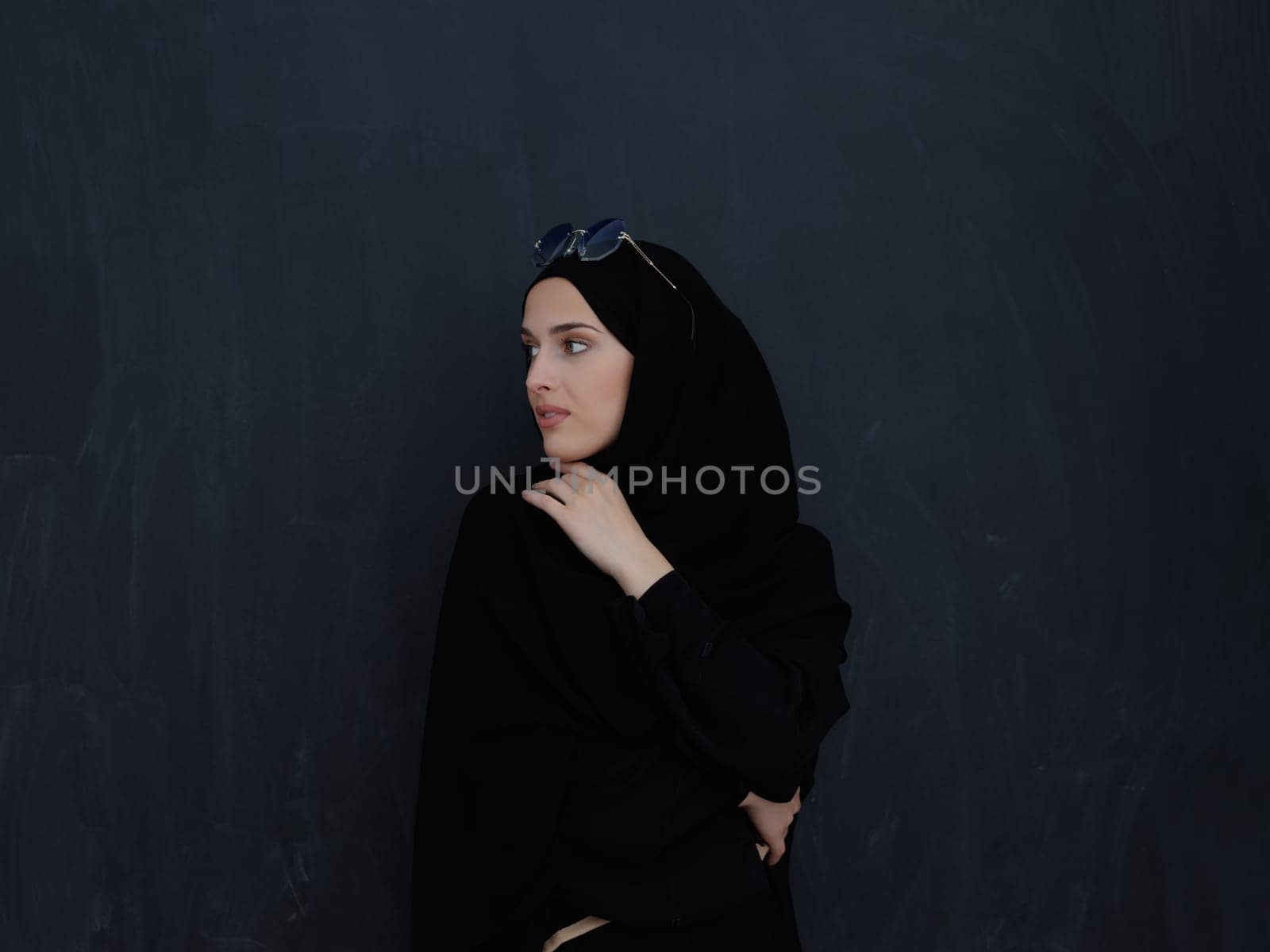 Young muslim in traditional clothes or abaya and sunglasses posing in front of black chalkboard. Arab woman representing modern arabic lifestyle, islamic fashion and Ramadan kareem concept