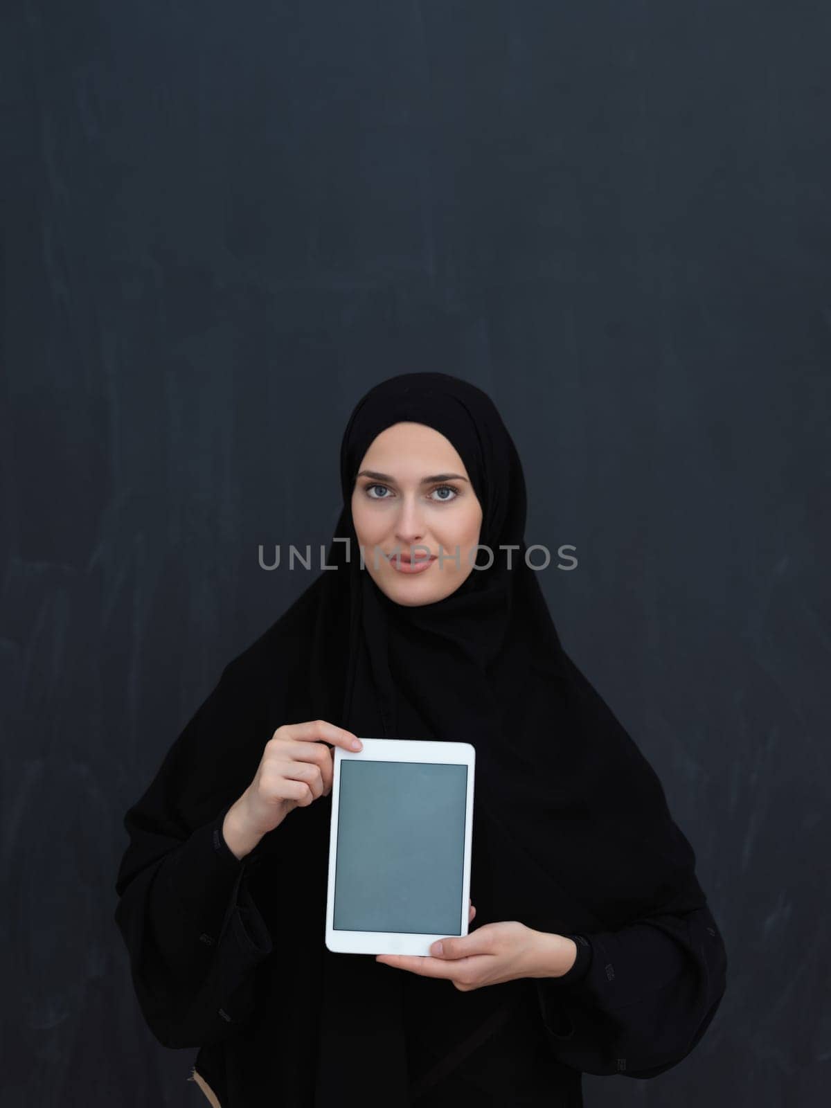 Young Arab businesswoman in traditional clothes or abaya and glasses showing tablet computer display in front of black chalkboard representing islam fashion and technology