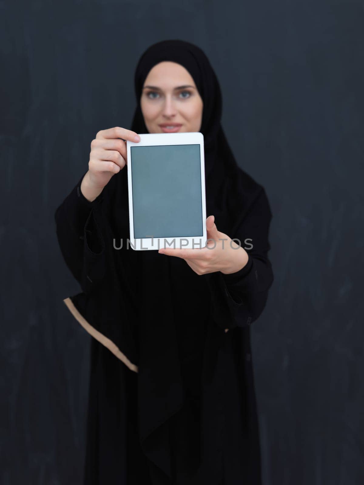 Young Arab businesswoman in traditional clothes or abaya and glasses showing tablet computer display in front of black chalkboard representing islam fashion and technology