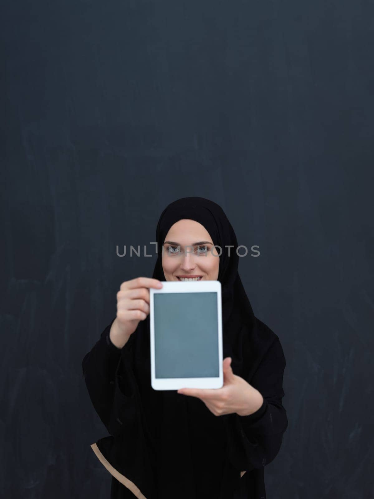 Young Arab businesswoman in traditional clothes or abaya and glasses showing tablet computer display in front of black chalkboard representing islam fashion and technology