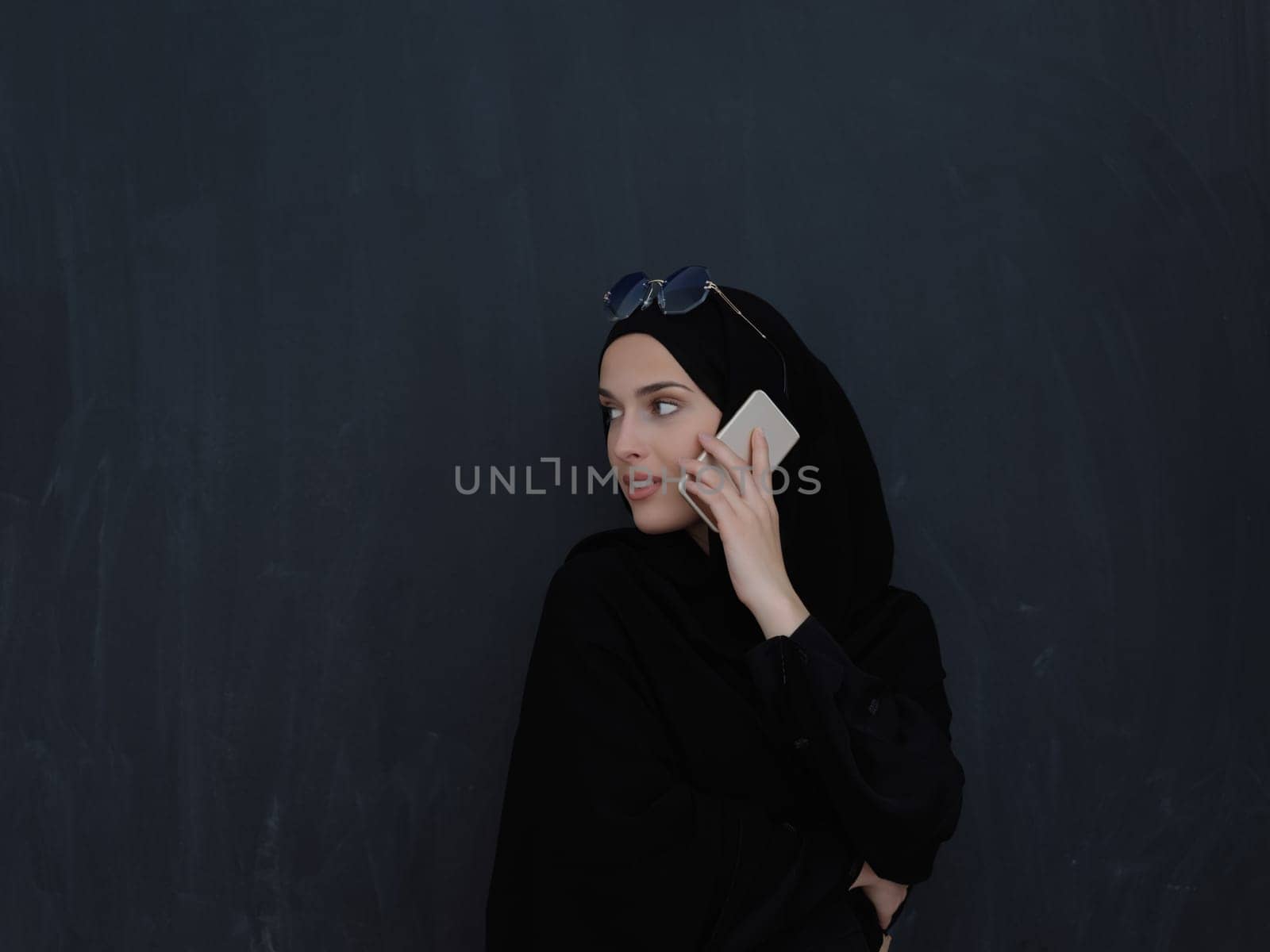 Young muslim businesswoman in traditional clothes or abaya talking on the smartphone. Arab woman in front of black chalkboard and representing techology, islamic fashion and Ramadan kareem concept