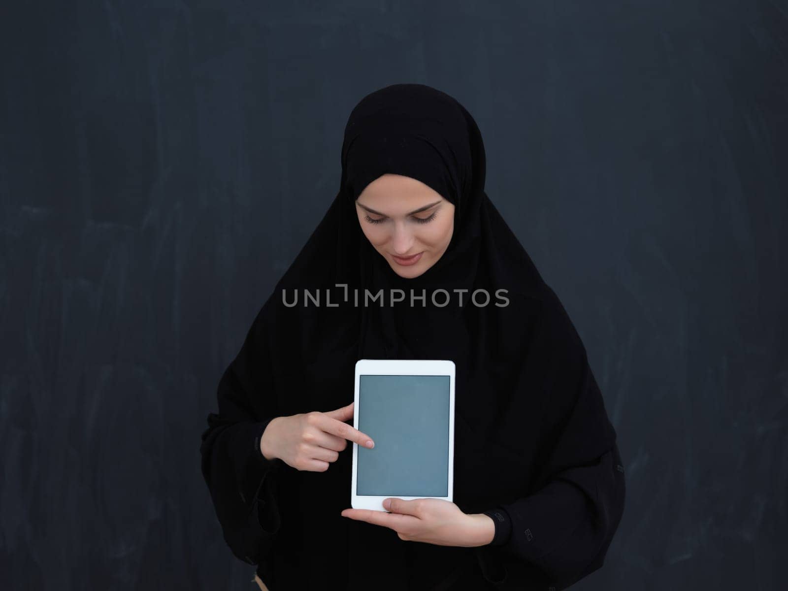Young Arab businesswoman in traditional clothes or abaya and glasses showing tablet computer display in front of black chalkboard representing islam fashion and technology