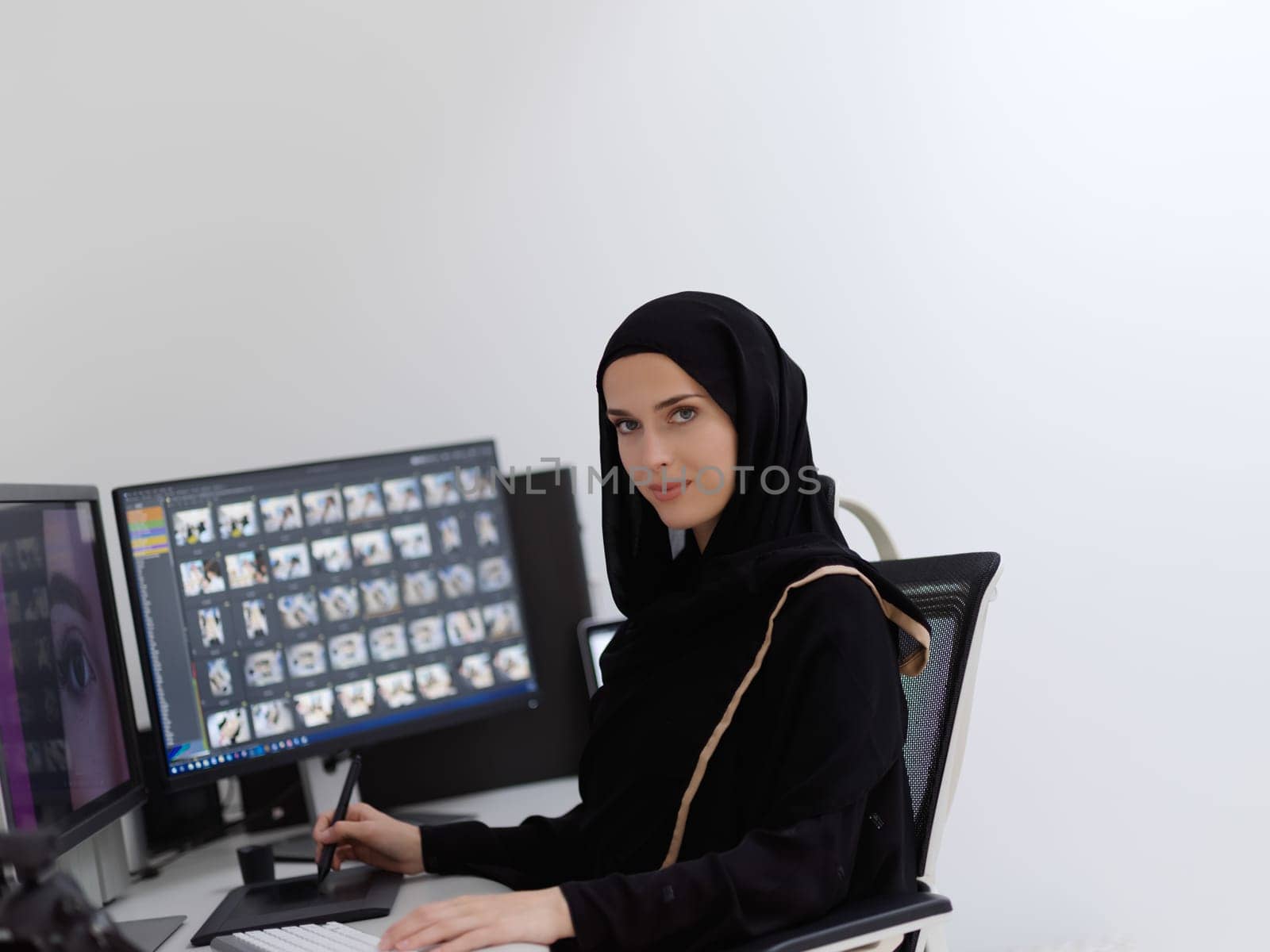 Muslim female graphic designer working on computer using graphic tablet and two monitors. Girl wearing hijab editing photos in the office