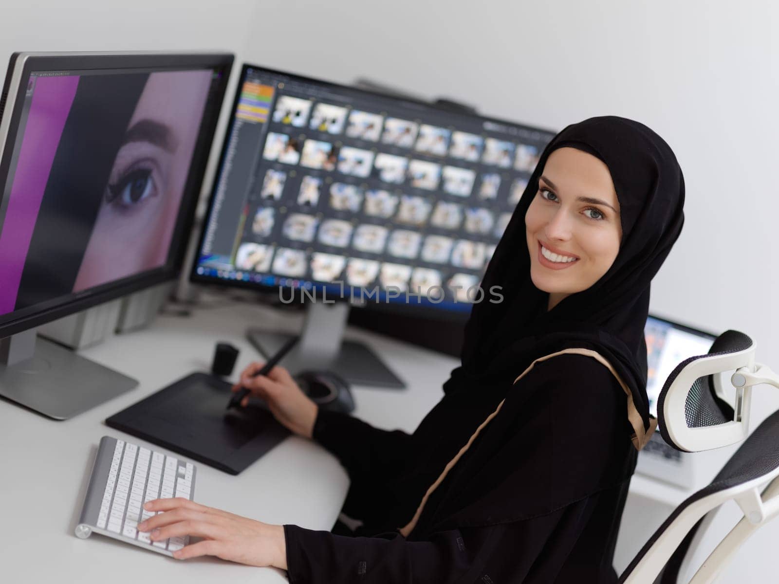 Muslim female graphic designer working on computer using graphic tablet and two monitors. Girl wearing hijab editing photos in the office