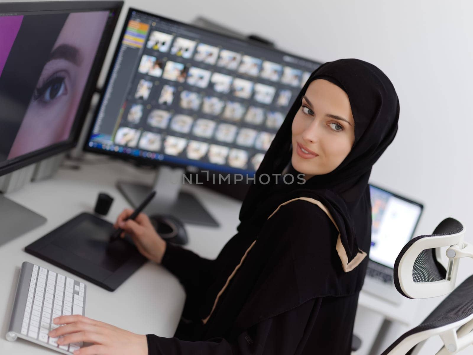 Muslim female graphic designer working on computer using graphic tablet and two monitors. Girl wearing hijab editing photos in the office