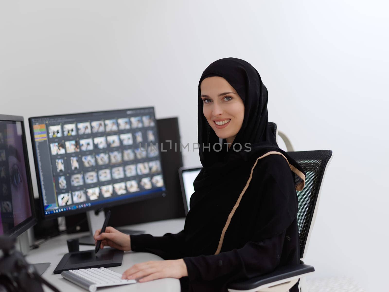 Muslim female graphic designer working on computer using graphic tablet and two monitors. Girl wearing hijab editing photos in the office