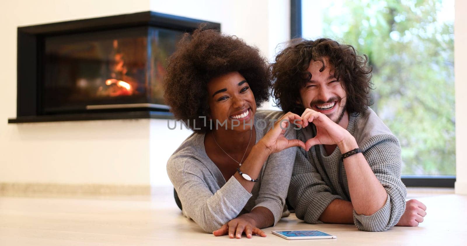 beautiful young multiethnic couple with tablet computer showing a heart with their hands on the floor in front of fireplace at autumn day