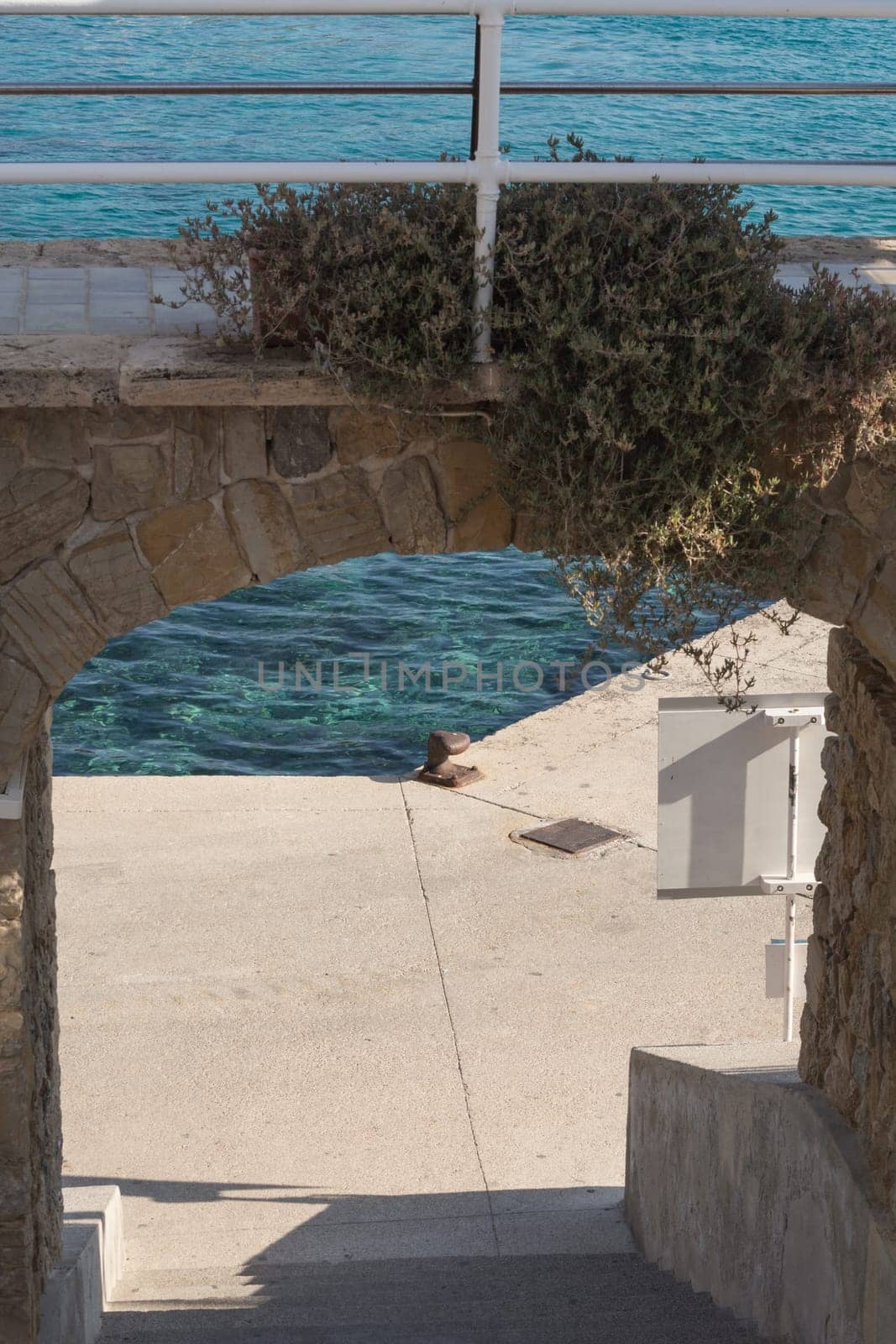 A rusty bollard on a quay wall with a rusty chain and in the background the water in the harbor on the Mediterranean Sea.