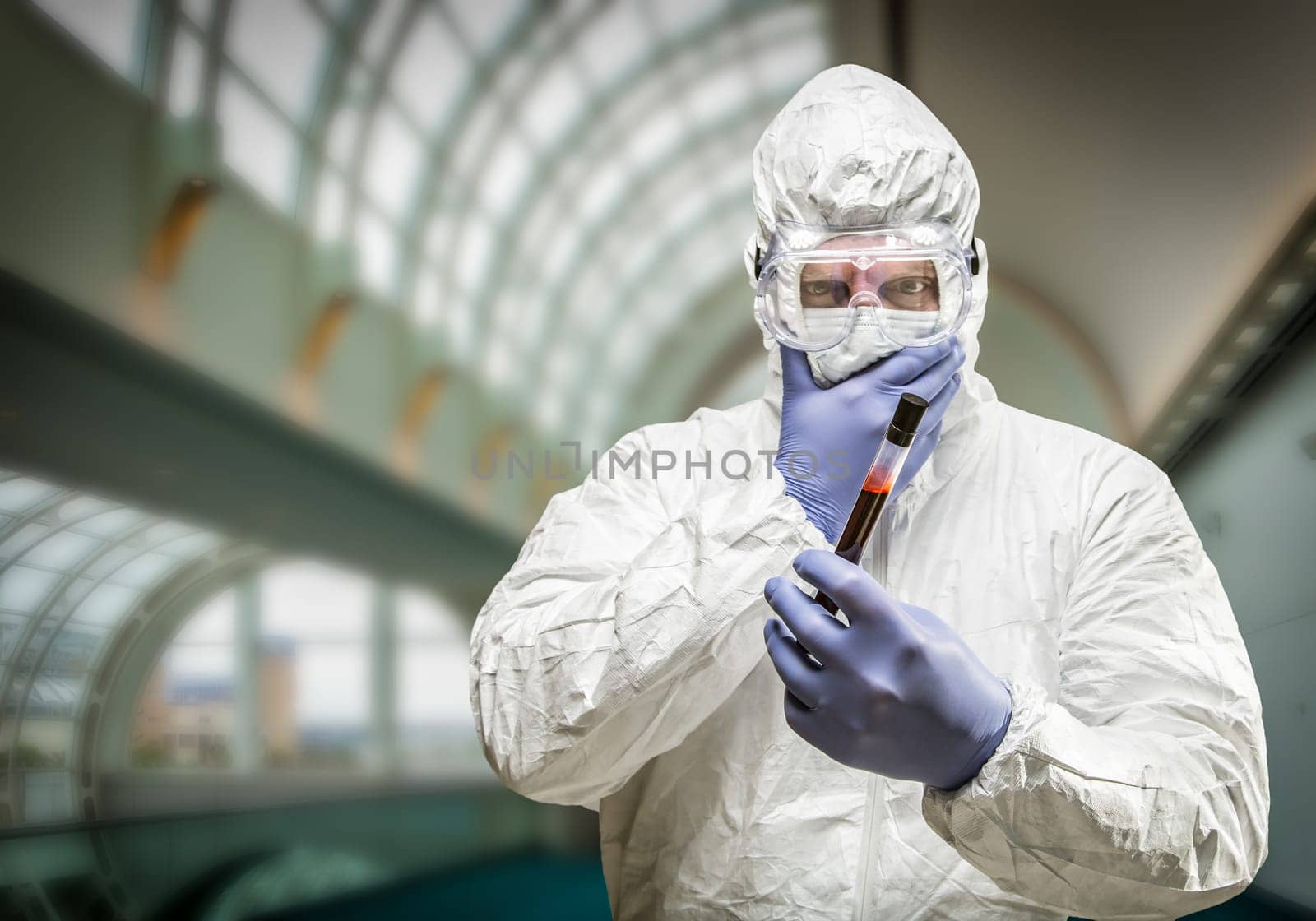 Man Wearing HAZMAT Protective Clothing Holding Test Tube Filled With Blood Inside Building.