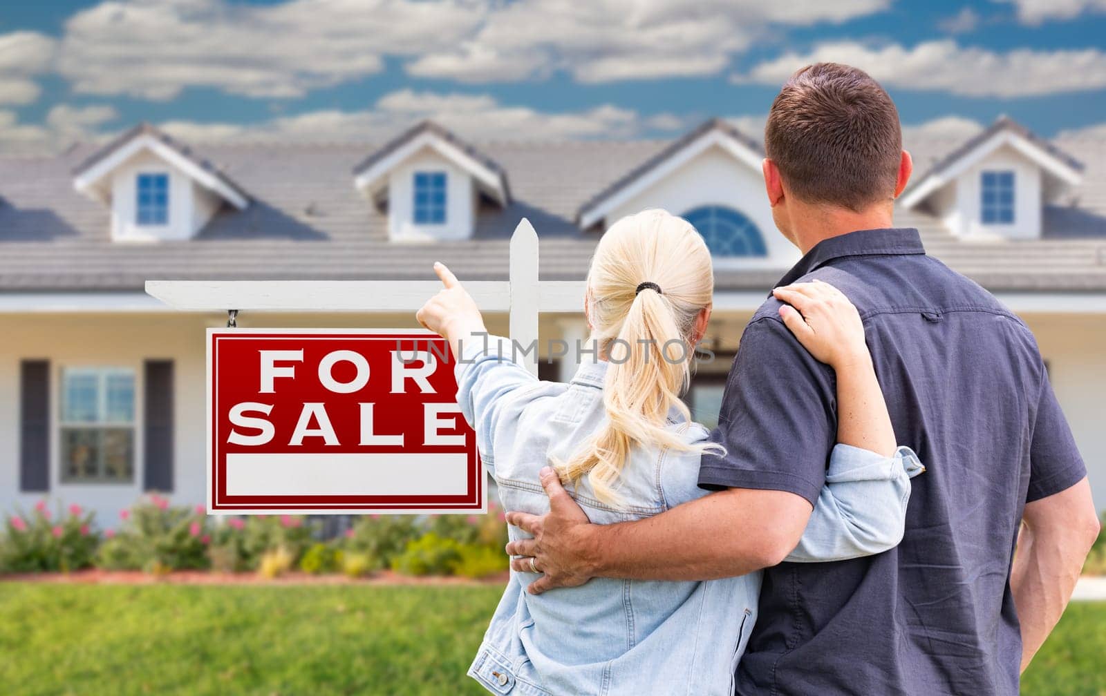 Young Adult Couple Facing and Pointing to Front of For Sale Real Estate Sign and House.