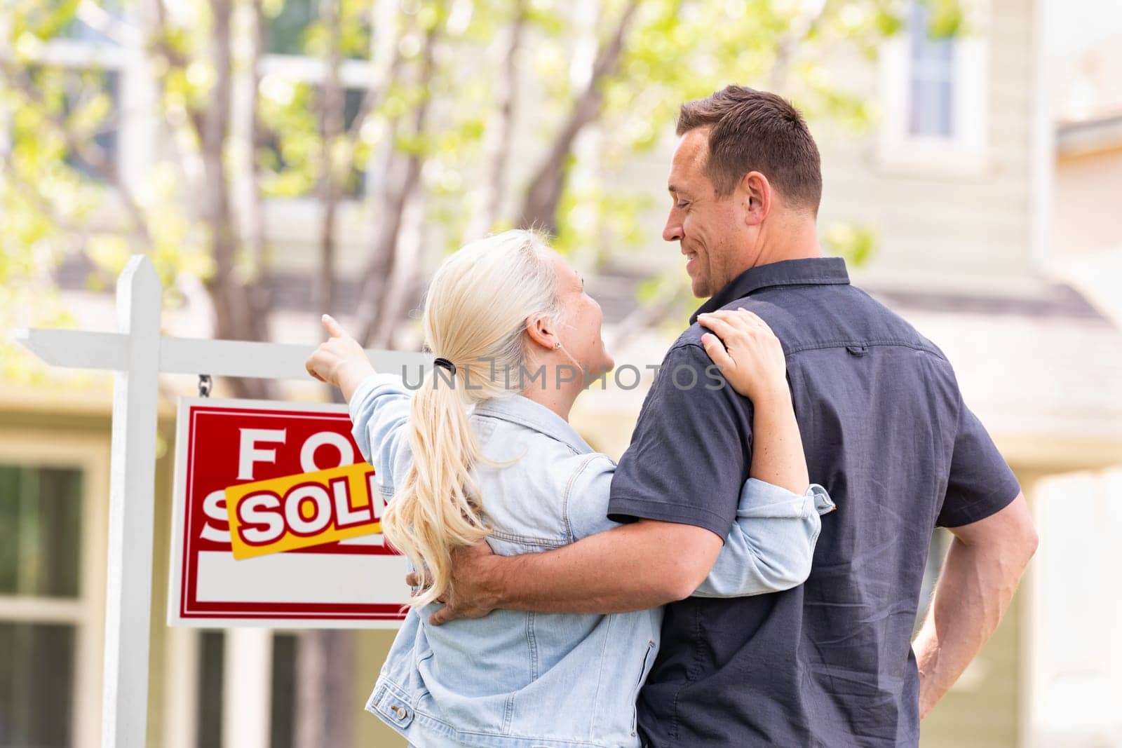 Caucasian Couple Facing and Pointing to Front of Sold Real Estate Sign and House.