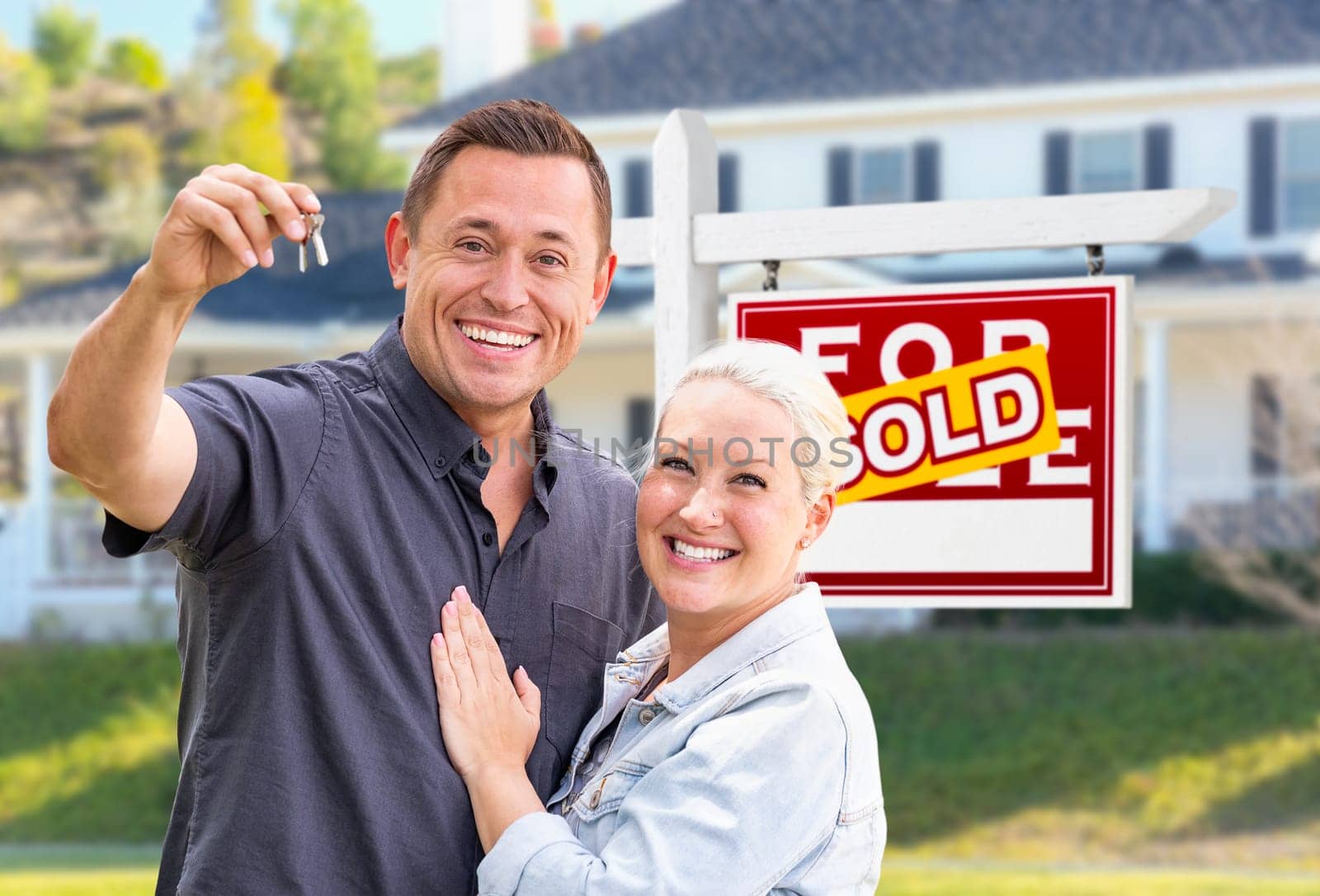 Young Adult Couple With House Keys In Front of Home and Sold For Sale Real Estate Sign.