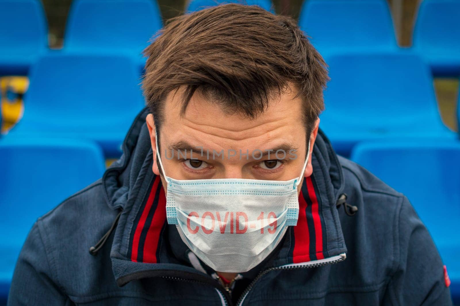 Man in surgical mask sits alone in stadium. COVID-19 Pandemic Coronavirus. Man in city stadium wearing face mask protective for spreading of Coronavirus Disease 2019. Portrait of man with surgical mask on face against virus.