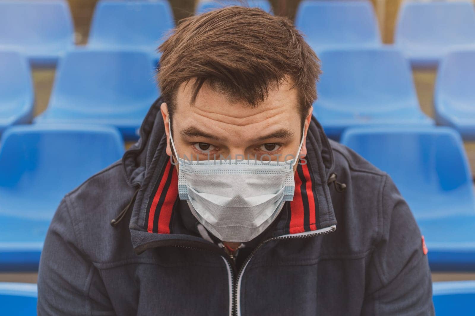 Man in surgical mask sits alone in stadium. COVID-19 Pandemic Coronavirus. Man in city stadium wearing face mask protective for spreading of Coronavirus Disease 2019. Portrait of man with surgical mask on face against virus.