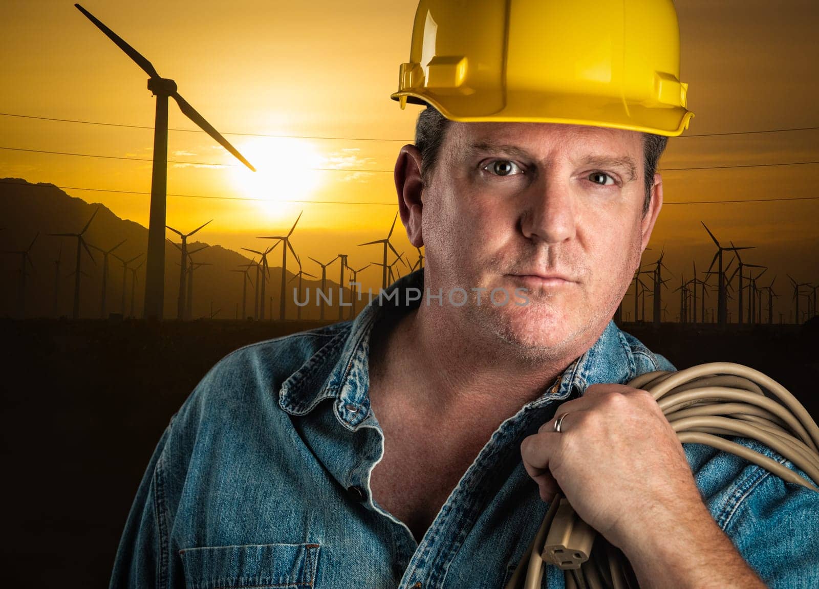 Serious Contractor in Hard Hat Holding Extension Cord Outdoors Near Wind Turbines.