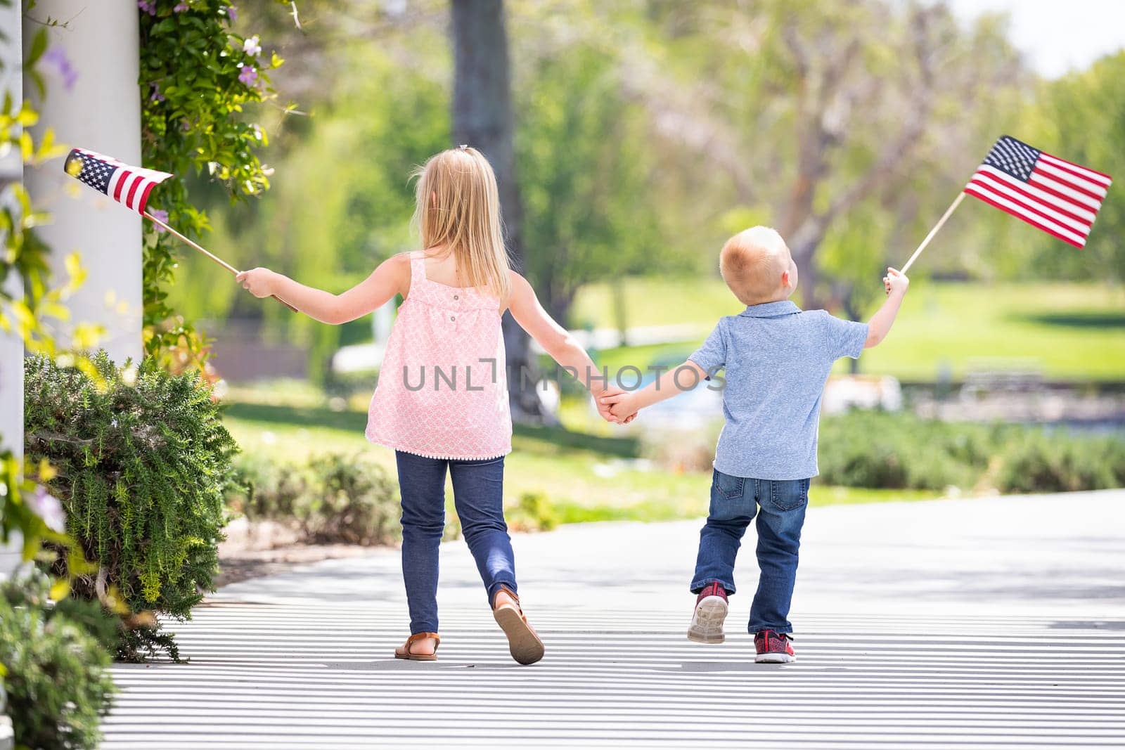 Young Sister and Brother Holding Hands and Waving American Flags At The Park.