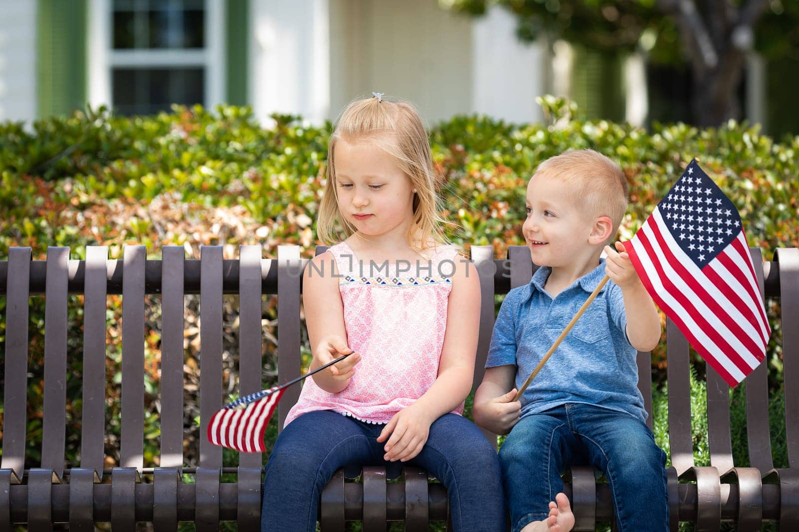 Young Sister and Brother Comparing Each Others American Flag Size On The Bench At The Park.
