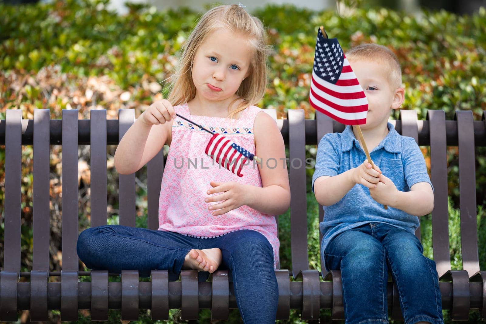 Young Sister and Brother Comparing Each Others American Flag Size On The Bench At The Park.