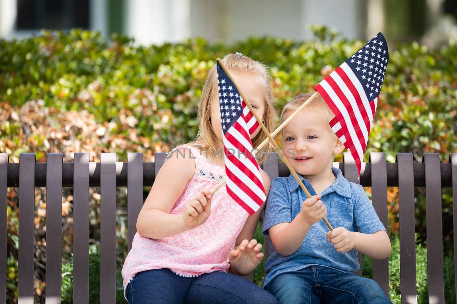 Young Sister and Brother Waving American Flags On The Bench At The Park.