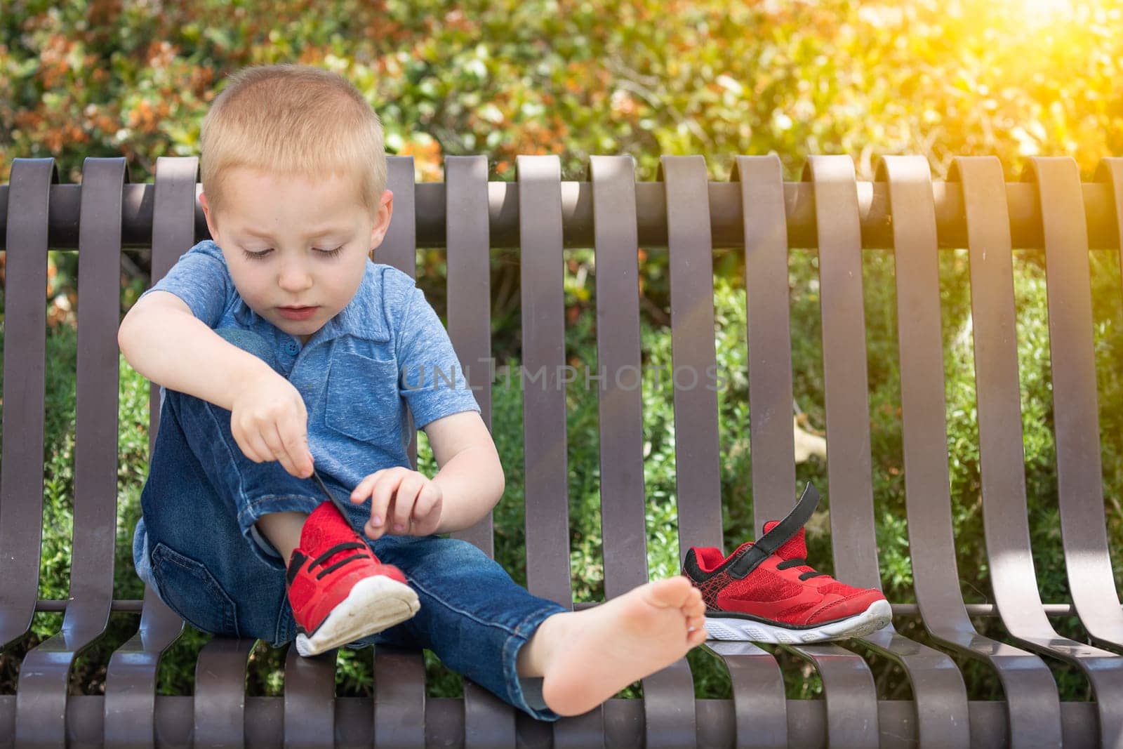Young Boy Sitting On A Bench Putting On His Shoes At The Park.