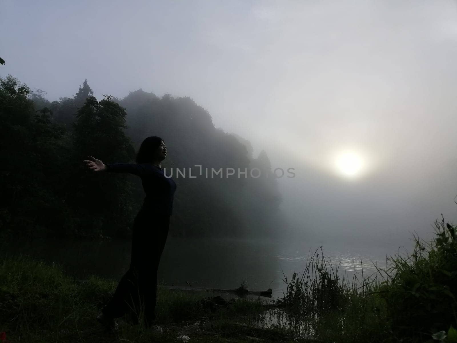 Young asian woman standing alone on lake enjoying peaceful moment of sunrise in gray and cloudy sky in the early chilly morning.  Concepts of winner, freedom, happiness.