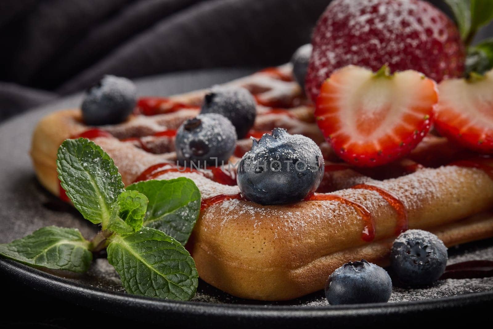 Delicious waffles with  powdered sugar and berries on ceramic black plate on grey background. Sweet meal. Dessert. Serving food