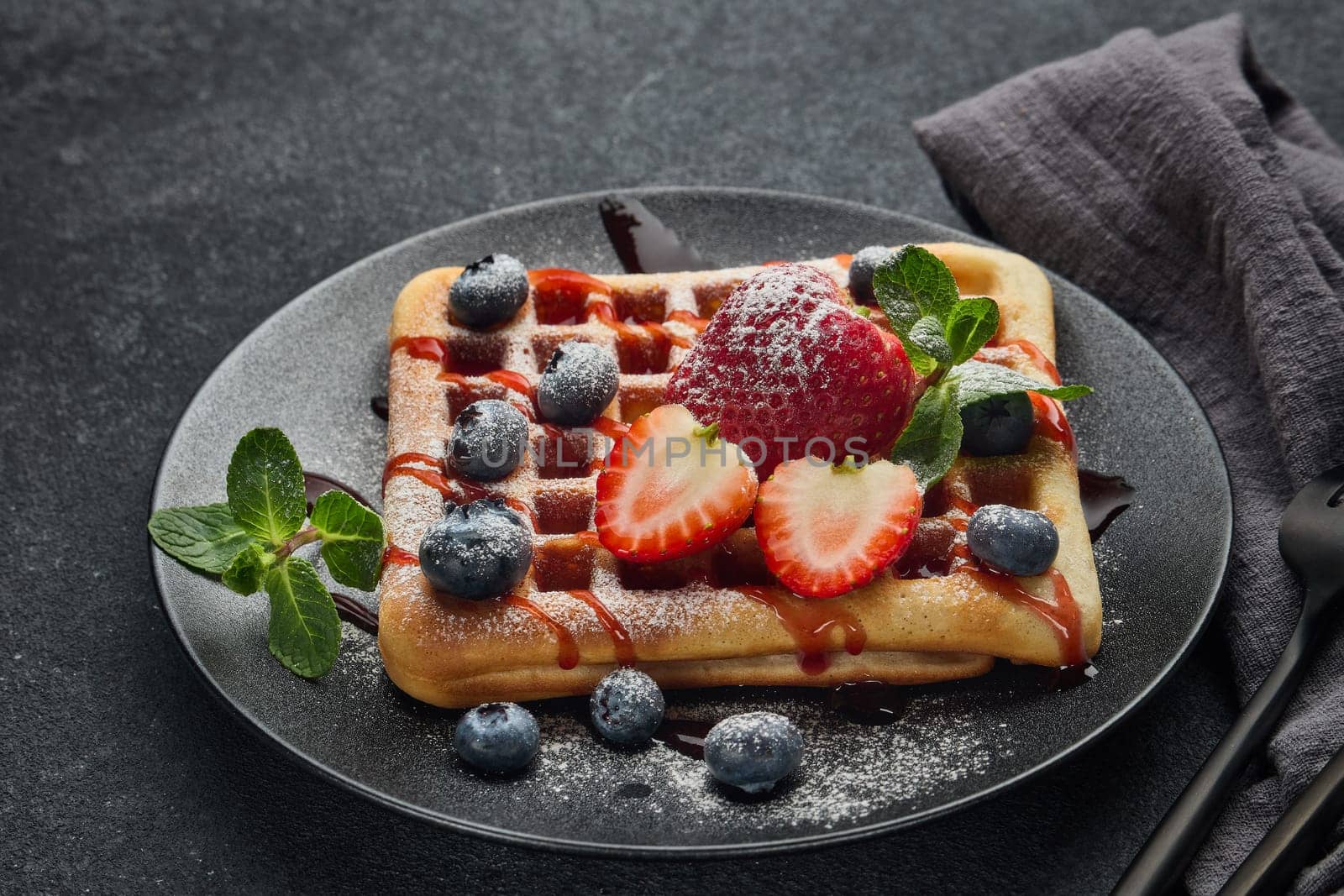 Delicious waffles with  powdered sugar and berries on ceramic black plate on grey background. Sweet meal. Dessert. Serving food