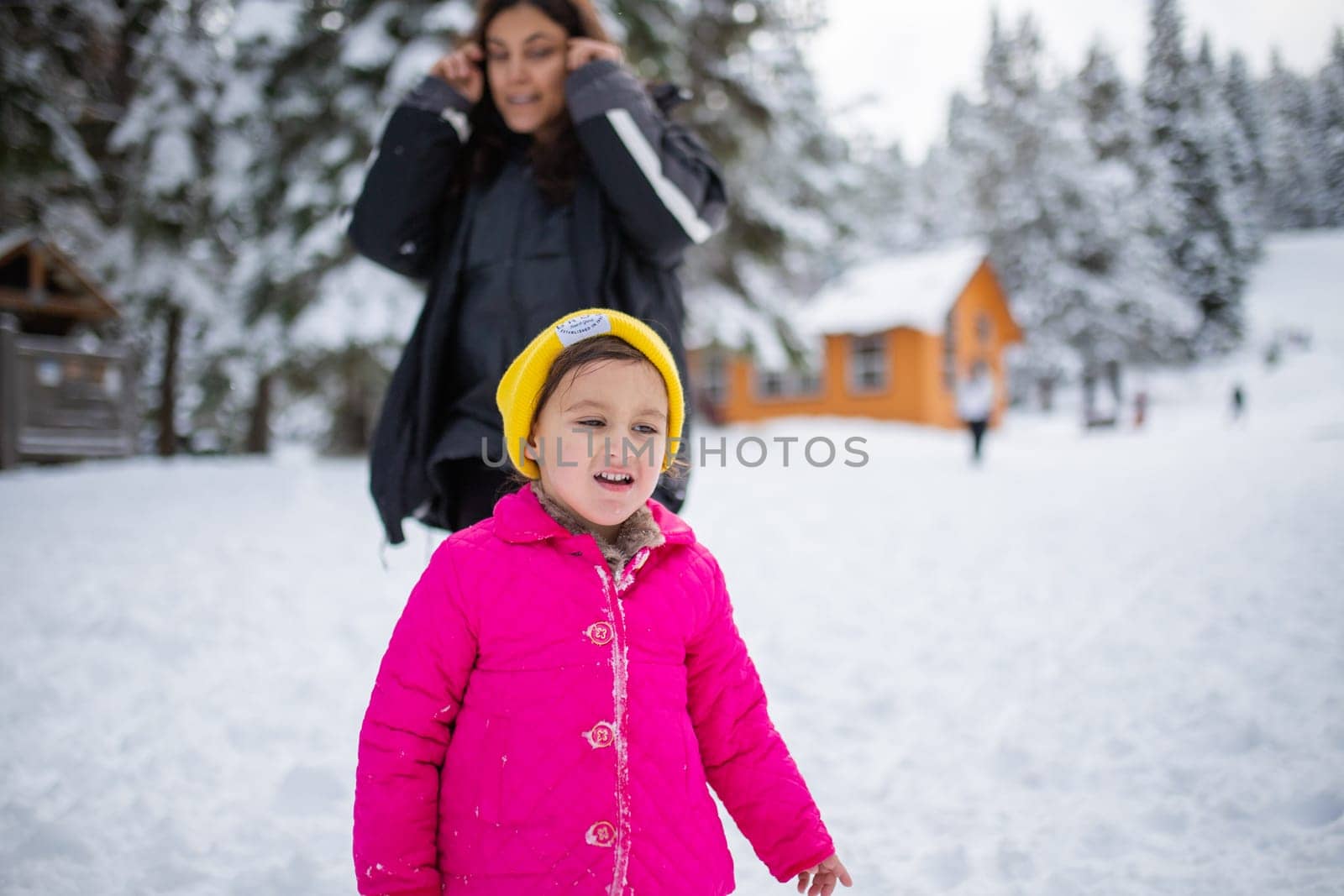 Happy little girl smiling with her mother behind her in snowy forest. Young daughter and mother having fun on snowy day. Adventurous winter holiday