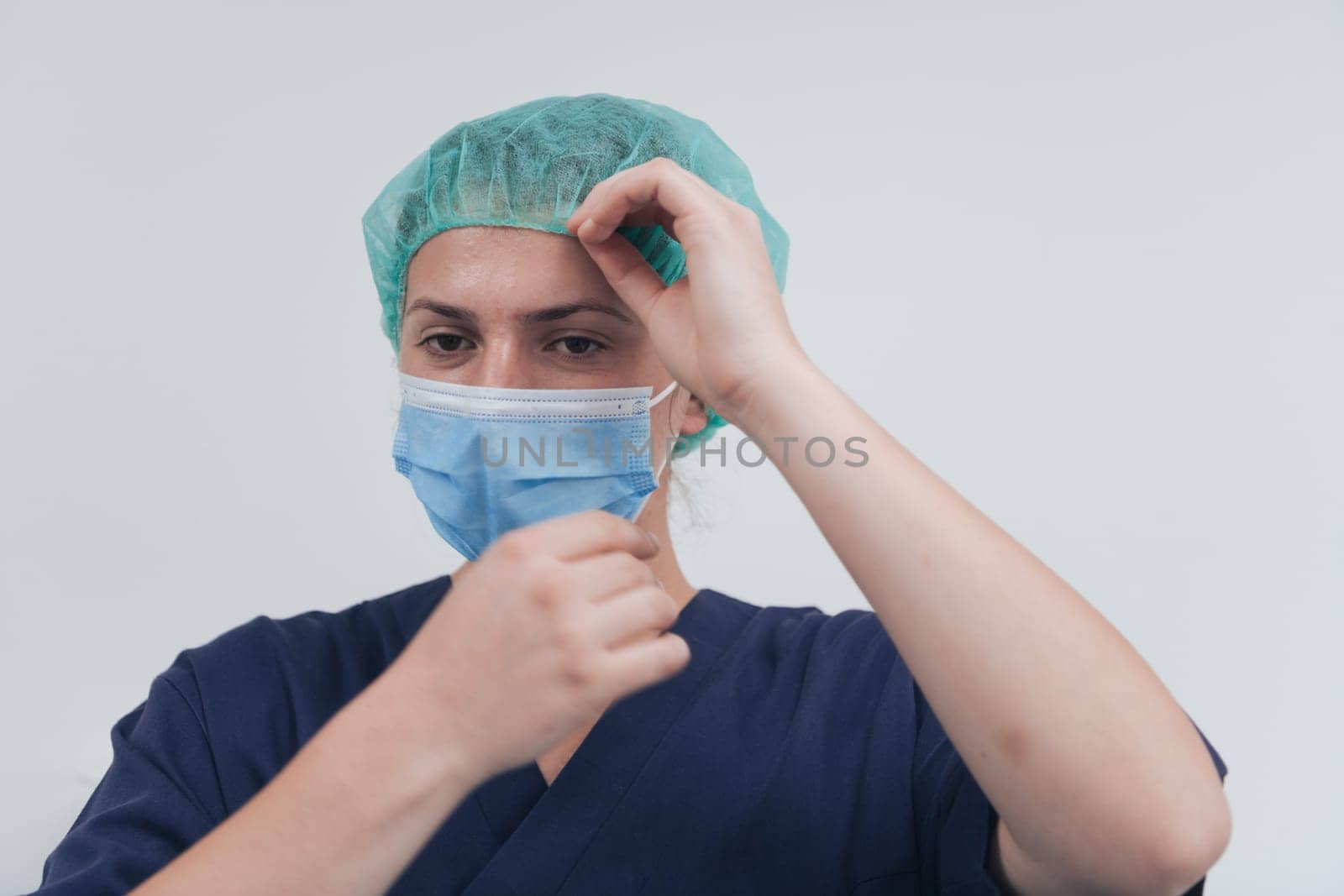 Close up of tired female doctor or scientist with a medical mask and surgical cap over grey background. Authentic real medical people hero in white in coronavirus pandemic is adjusting mask.