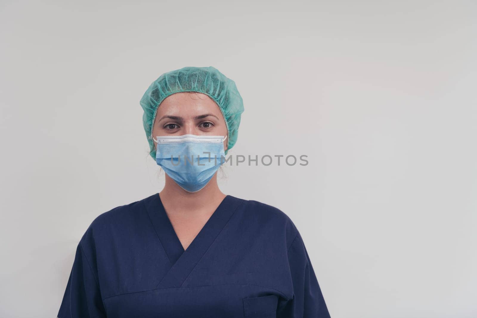 Close up of tired female doctor or scientist with a medical mask and surgical cap over grey background. Authentic real medical people hero in white in coronavirus pandemic is adjusting mask.
